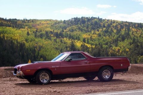 1972 Ford Ranchero - A maroon 1972 Ford Ranchero pickup truck is seen from the side, parked on a dirt road with a backdrop of trees in autumn colors.