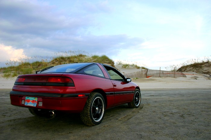 Red sport coupe rear 3/4 view parked on a beach with dunes behind