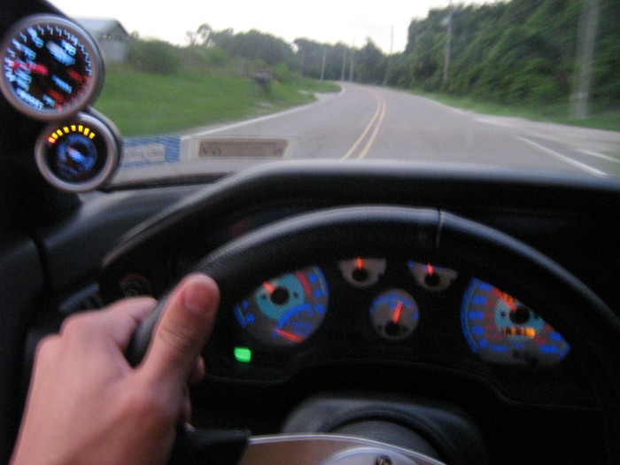 Driver POV of car interior, hand on wheel, blue gauges, and pillar-mounted aftermarket gauges on a rural road.
