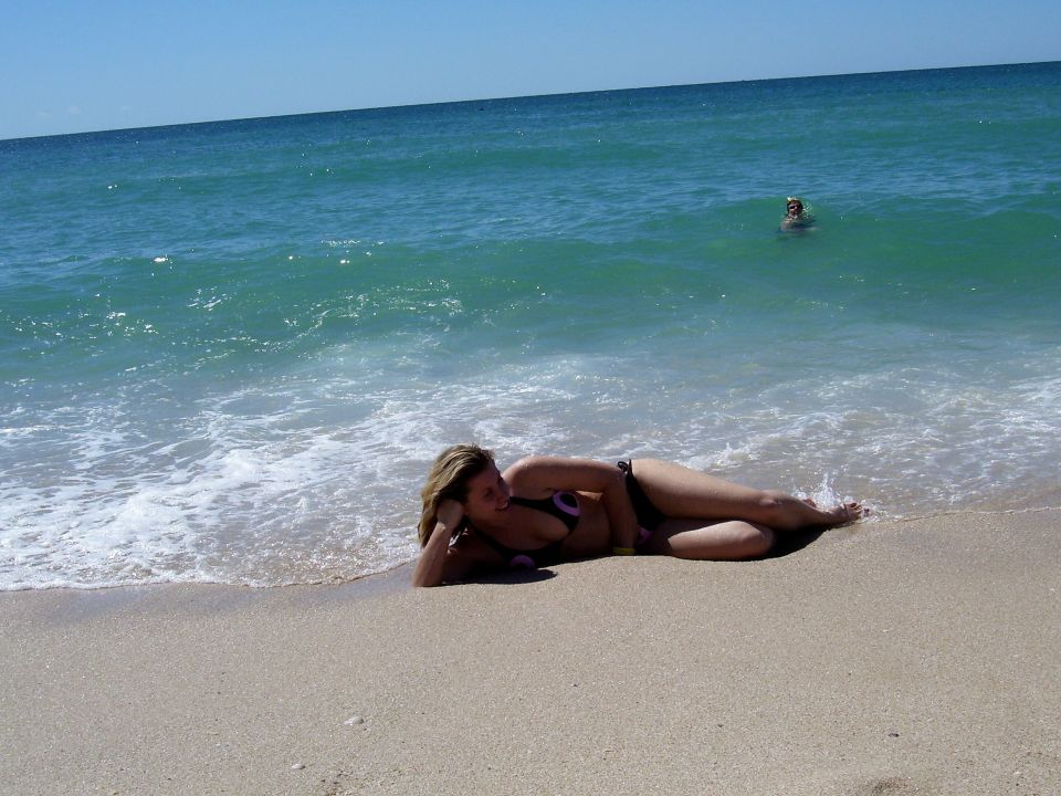 Woman in bikini lying on a sandy beach with waves washing ashore. Person swimming in the ocean in the background. SUPERMOTORS.NET logo in lower right