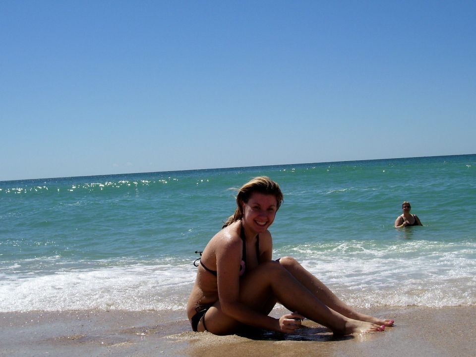 Woman in a bikini sits on a sandy beach with waves lapping at her legs