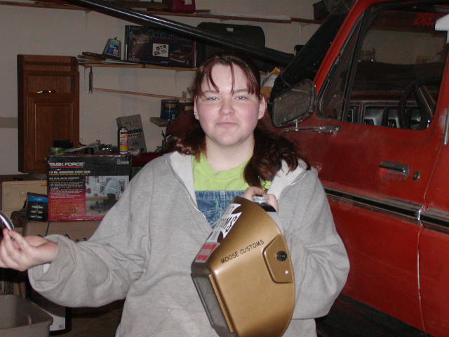 Person holding a gold welding mask with 'MOOSE CUSTOMS' text, standing in front of a red vehicle in a garage