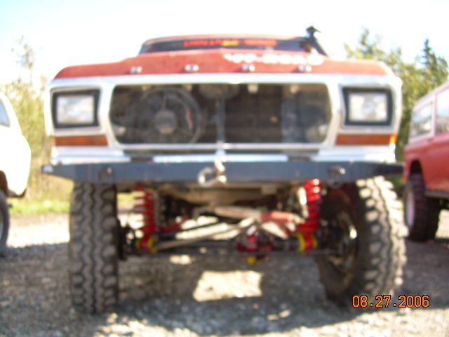 Front view of a red lifted truck with a fan visible in the grille. Another red vehicle is parked to the right