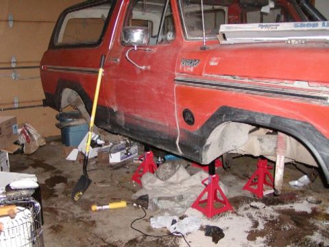 1978 Ford Bronco - A red 1978 Ford Bronco is elevated on jack stands inside a cluttered garage, undergoing maintenance.