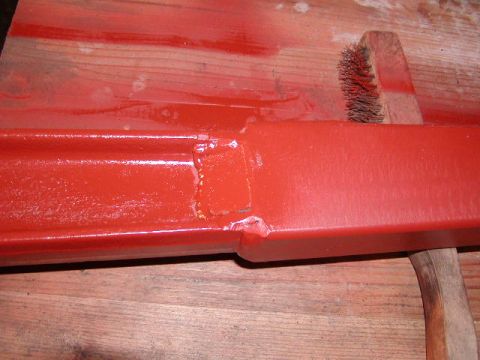 1978 Ford Bronco - A close-up view of a red painted metal beam with a wire brush and part of a wooden handle.