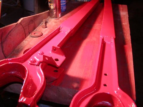 1978 Ford Bronco - Two metal components, freshly painted bright red, sit on a wooden workbench.