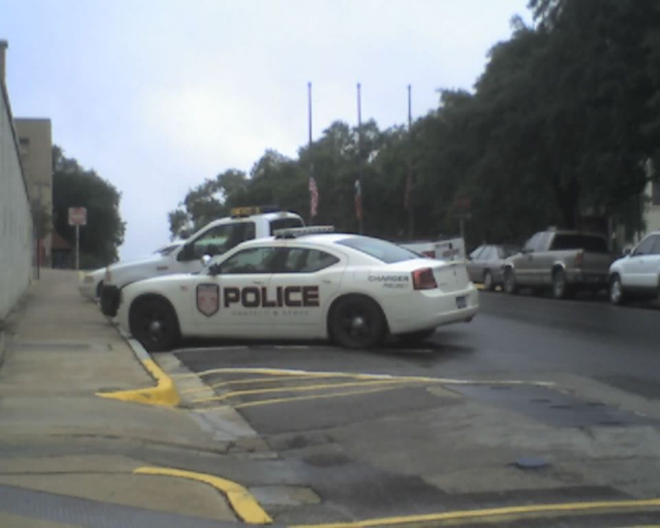 White police car parked on street, text on door says 'POLICE'. Another police vehicle and two trucks are in the background