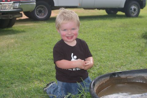 1998 Jeep TJ - A young child kneels on the grass, smiling at the camera, with two trucks in the background.