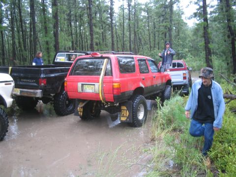 1990 Ford Bronco - Multiple off-road vehicles are stuck in deep mud in a wooded area.