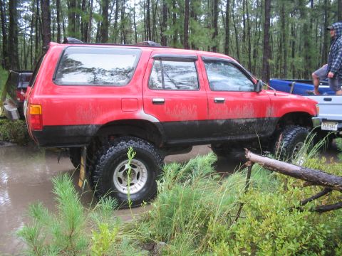 1990 Ford Bronco - A red SUV navigates through a muddy, wooded area with a pickup truck nearby.