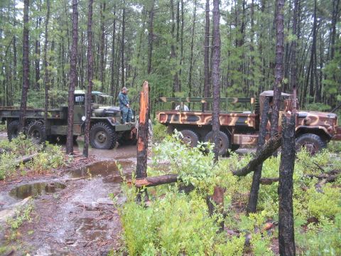 1990 Ford Bronco - Two military trucks, one green and one camouflage, are parked in a muddy forest with trees all around.