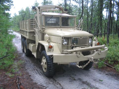 1990 Ford Bronco - A tan military truck drives down a dirt road surrounded by trees.