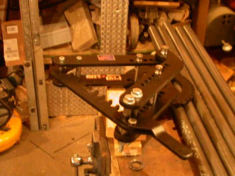 1990 Ford Bronco - A manual tubing bender with a black triangular frame, bolted-on degree ring and long handle mounted at a workbench, with lengths of round s…