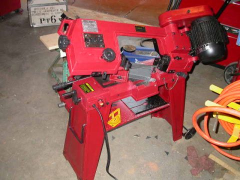 1990 Ford Bronco - A red horizontal metal-cutting band saw with a black electric motor sitting on its stand on a garage floor, coiled orange air hose and a re…