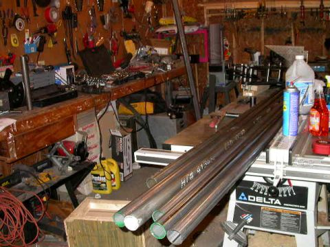 1990 Ford Bronco - Bundle of long round steel tubing laid across a Delta table saw in a home workshop, with green-painted cut ends and chalk markings on the t…