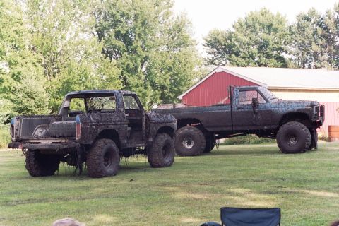 1979 Ford Bronco - Two heavily mud-caked Ford vehicles, a Bronco and a pickup truck, sit on a grassy field next to a red barn.