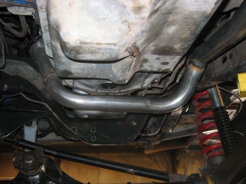 1992 Ford Bronco - Underside view of a vehicle showing a bare metal exhaust pipe or crossover routed beneath the transmission pan, with a red coil spring and…