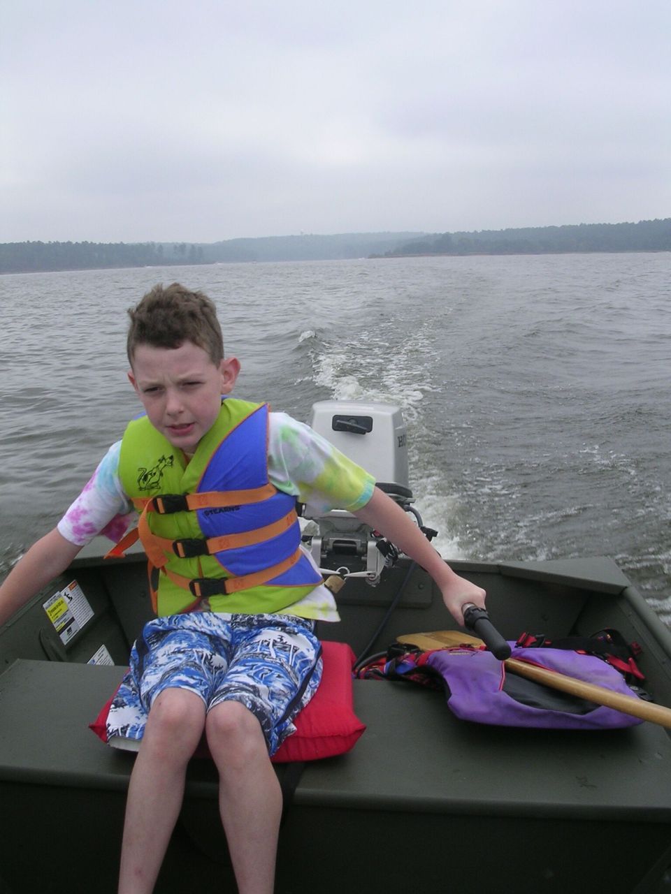 Boy in colorful life jacket seated in aluminum boat on a gray lake