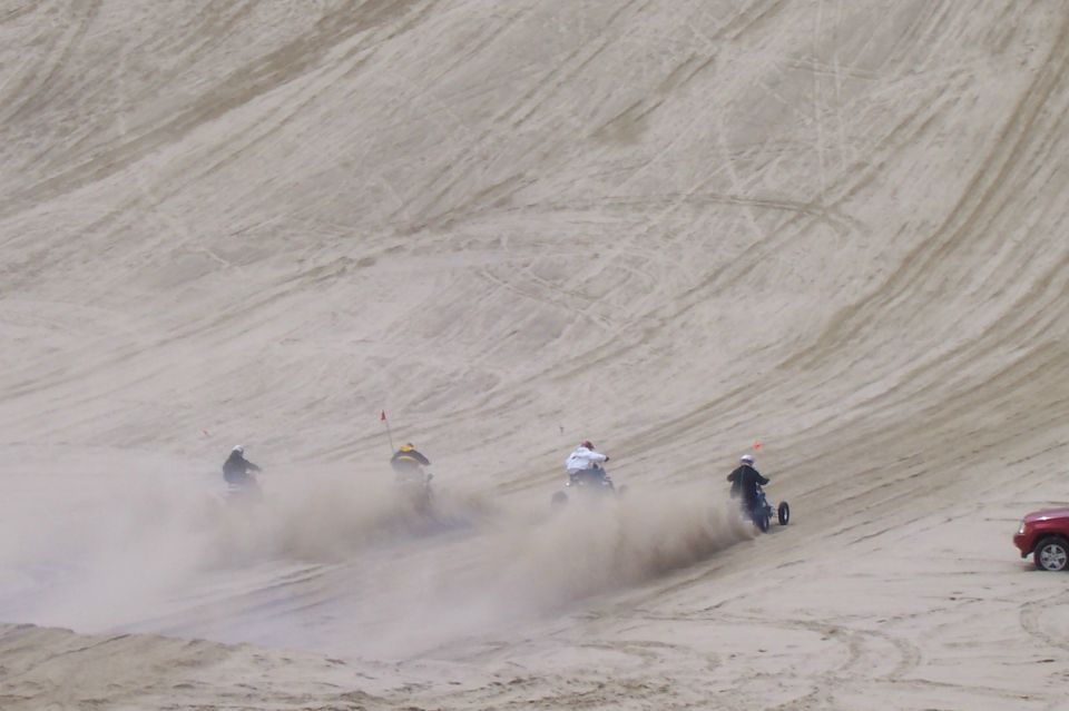Four three-wheeled vehicles kick up dust as they ride down sand dunes. A red SUV is parked on the right