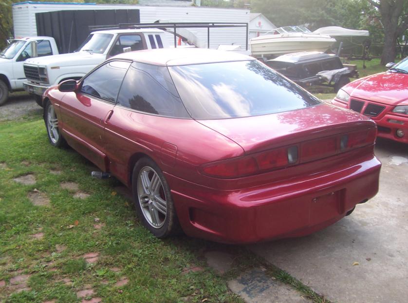 Red coupe parked in a yard with a white truck and boats in background. Supermotors.net logo visible