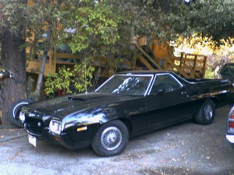 1972 Ford Ranchero - black early-1970s Ford Ranchero car-pickup with wire wheels and hood scoop parked under trees near a wooden staircase.