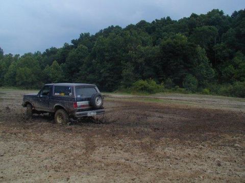 1988 Ford Bronco - A dark blue Ford Bronco navigates a muddy off-road course, kicking up dirt as it goes.