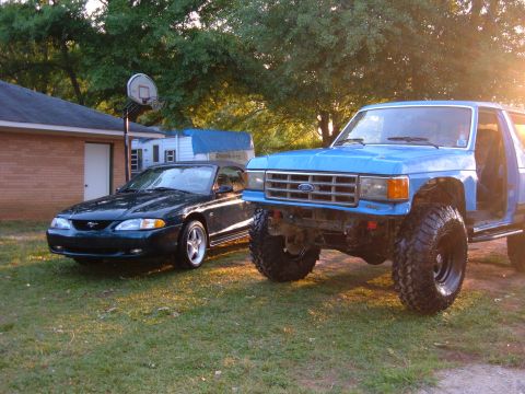 1987 Ford Bronco - Two Ford vehicles, a lifted truck and a Mustang, are parked outside a house with a trailer.