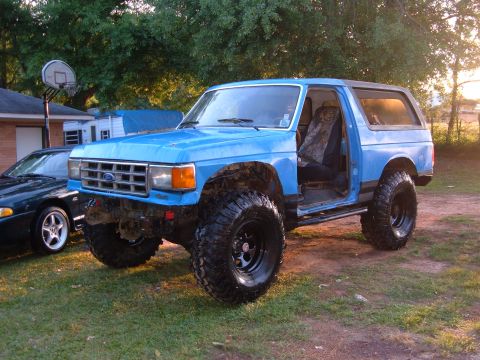 1987 Ford Bronco - This lifted 1987 Ford Bronco is parked on a grassy area with a dark sedan nearby and trees in the background.