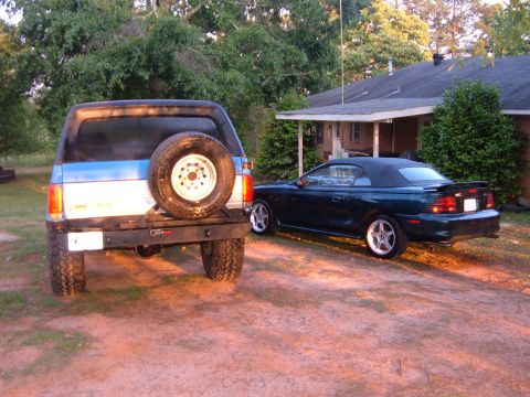 1987 Ford Bronco - A Ford Bronco and a Ford Mustang convertible are parked in a driveway next to a house.