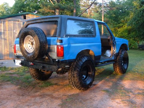 1987 Ford Bronco - This blue 1987 Ford Bronco features a rear-mounted spare tire, lifted suspension, and aggressive off-road tires.