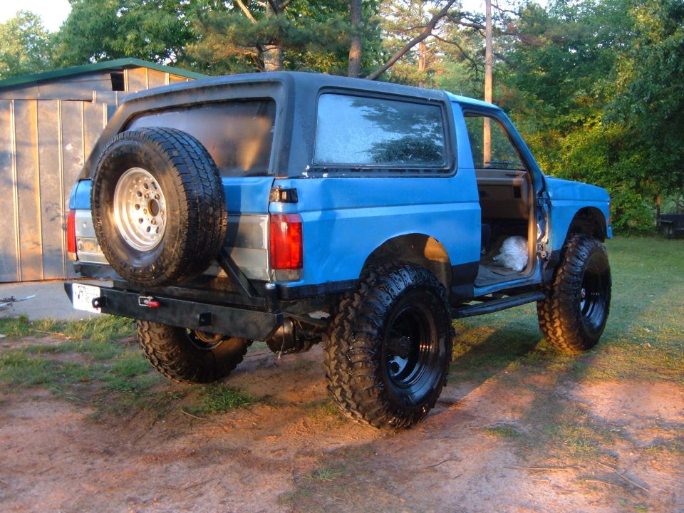 Blue 1987 Ford Bronco with spare tire mounted on rear bumper, lifted suspension, and off-road tires