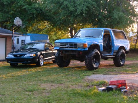 1987 Ford Bronco - Two Ford vehicles, a lifted Bronco and a Mustang, sit in a yard with a trailer and house in the background.