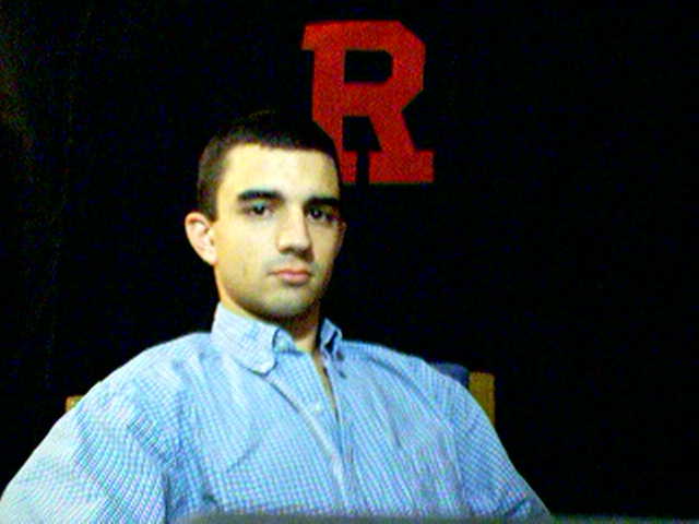 Young man in a blue checked shirt seated in front of a black backdrop with a red letter R