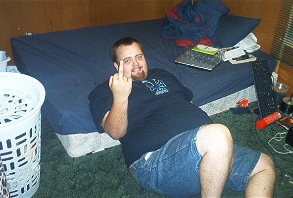 Man in a t-shirt and denim shorts lies on a bed, giving the middle finger