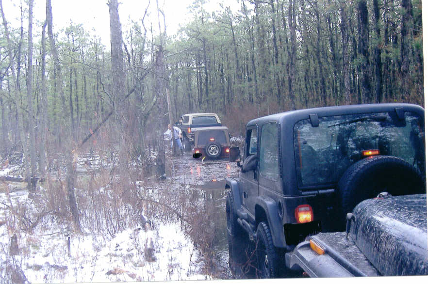 Black Jeep Wrangler rear view, driving on a muddy forest road with snow patches