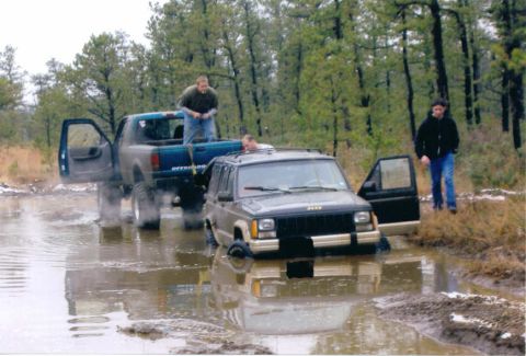 1997 Jeep TJ - Two vehicles are stuck in a muddy, wooded area. The Jeep Cherokee's front end is deep in the water.