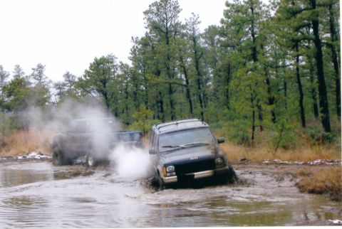 1997 Jeep TJ - Two SUVs navigate a muddy trail surrounded by pine trees, kicking up water and dirt.