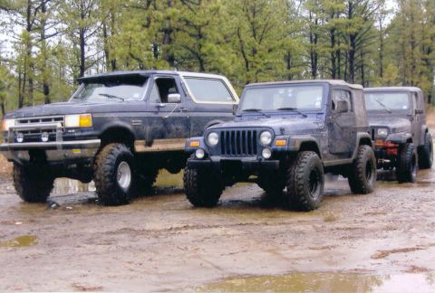 1997 Jeep TJ - Three off-road vehicles, including a blue Jeep TJ, are parked on a muddy track surrounded by trees.
