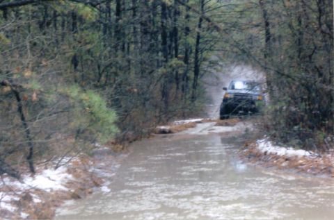 1997 Jeep TJ - A dark SUV navigates a wet and muddy path through a dense forest.