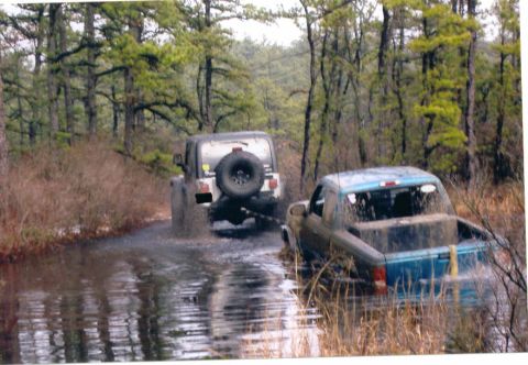 1997 Jeep TJ - Two off-road vehicles, a 1997 Jeep TJ and a Ford Ranger, navigate a muddy, water-filled trail in a wooded area.