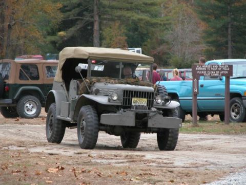 1997 Jeep TJ - A vintage military truck is parked on a dirt lot, with other vehicles and trees in the background.