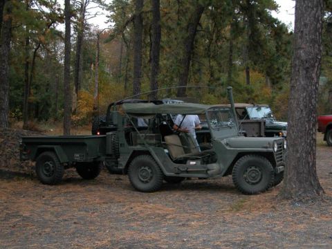 1997 Jeep TJ - A military jeep and trailer are parked in a forest clearing, with another vehicle visible in the background.