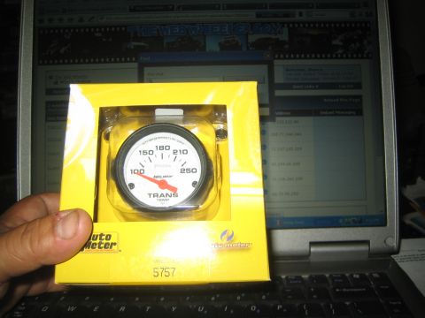 1990 Ford Bronco - hand holds a boxed AutoMeter transmission temperature gauge in front of a laptop displaying a SuperMotors web page.