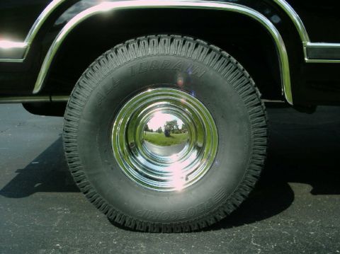 1984 Ford Bronco - A close-up shot focuses on the tire and chrome wheel of a black vehicle, reflecting the surrounding environment.
