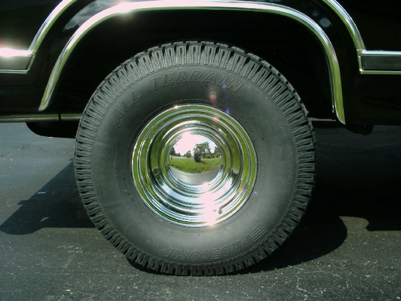 Close-up of a black vehicle's tire and chrome wheel. The tire has 'ALL TERRAIN' written on it. The wheel reflects a tree and a road