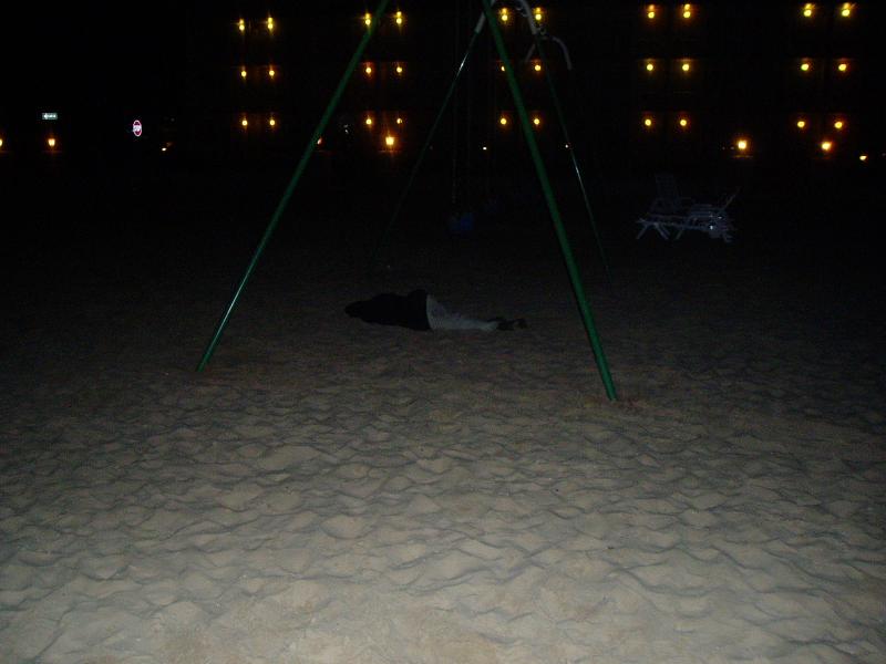 Person lying on sand under a swing set at night with lights in the background