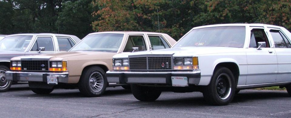 Three Ford Panther platform sedans parked in a row. The front one is white, the middle is tan, and the rear is dark