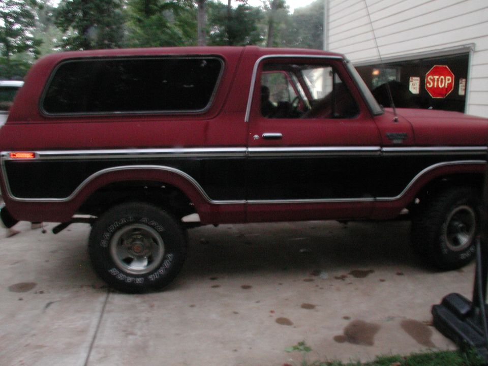 Red and black 1978 Ford Bronco side view, parked on concrete with a garage in the background