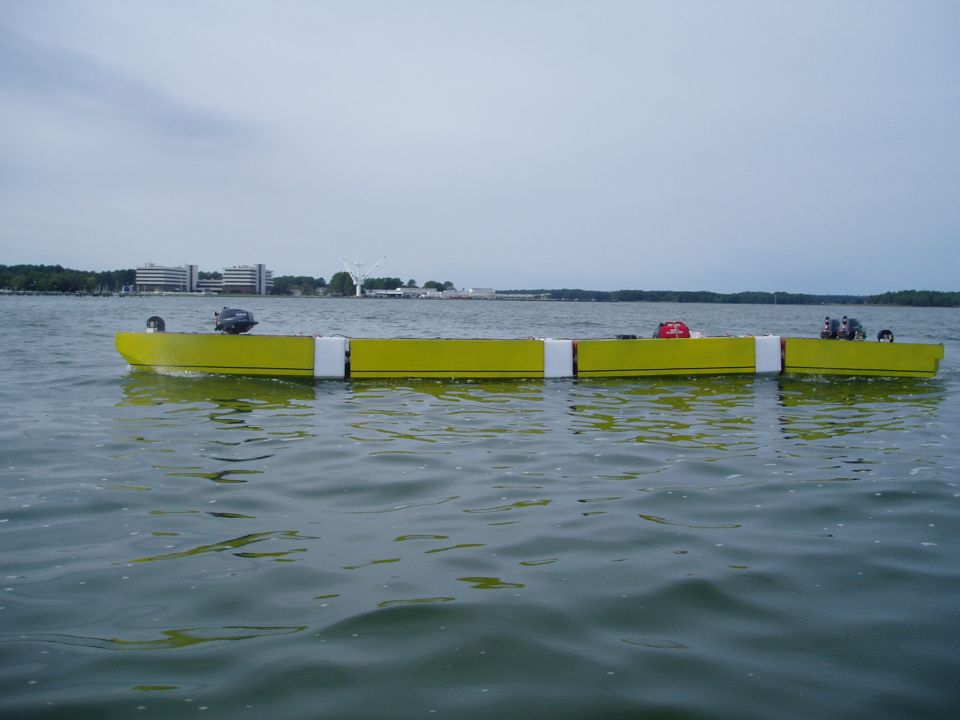 Yellow floating barrier with outboard motors on water, buildings in background