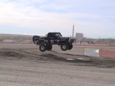 1990 Ford Bronco - black number 300 Ford pickup launches off a dirt jump on a desert course with an industrial plant in the background.
